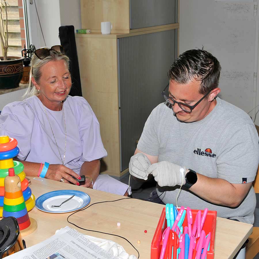 Eine Frau und ein junger Mann sitzen an einem Tisch. Der Mann hat silberfarbene Handschuhe an, an denen Kabel befestigt sind, und versucht, einen Faden in eine Nadel einzufädeln.