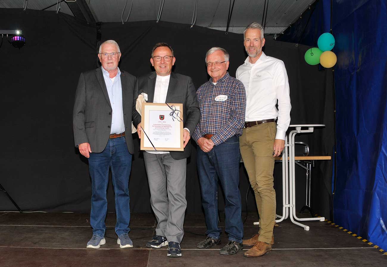 Kreispräsident Helmuth Ahrens, Michael Behrens, Gerhard Ramcke, Holger Rennemann stehen auf der Bühne, Herr Behrens zeigt die gerahmte Urkunde