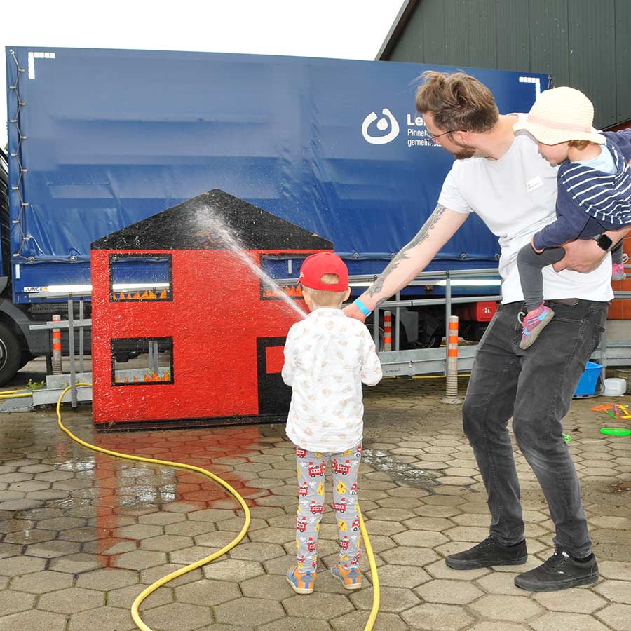 Ein kleiner Junge versucht, mit einem Wasserstrahl ein Loch in einem kleine Haus aus Holz zu treffen und wird von seinem Vater unterstützt.