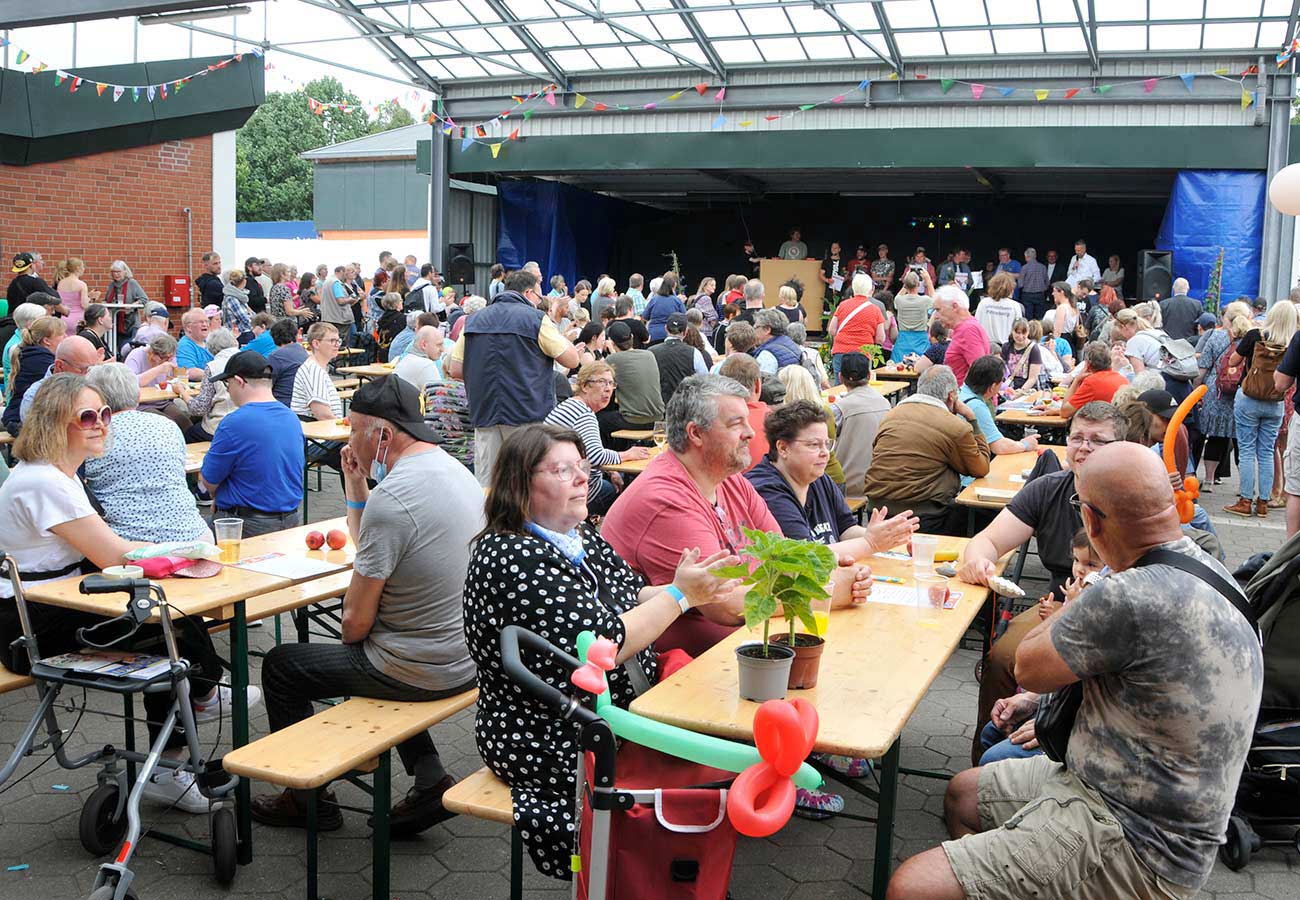 Viele Gäste sitzen auf dem Hof des Lebenshilfewerks Pinneberg auf Bänken und beobachten die Verleihung der Urkunden für die Jubilare.